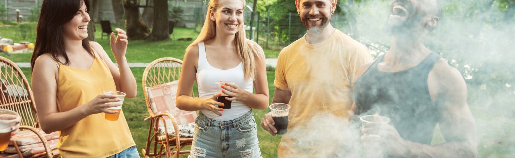 Group of happy friends having beer and barbecue party at sunny day. Resting together outdoor in a forest glade or backyard. Celebrating and relaxing, laughting. Summer lifestyle, friendship concept.