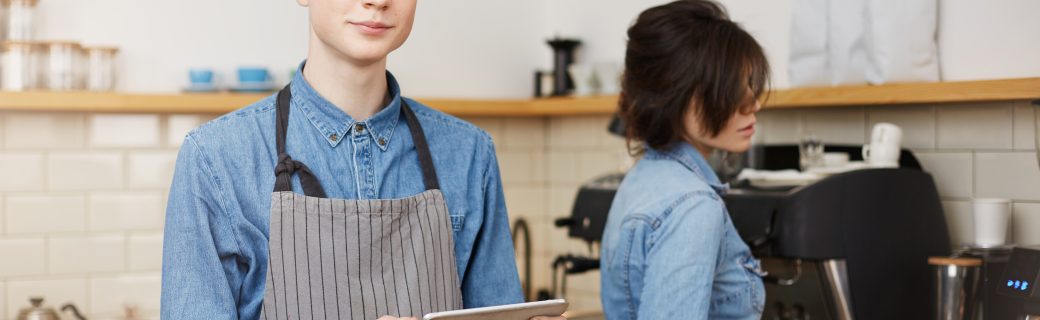 Male barista taking order, holding tab, female barista making preparing coffee using coffee tamper.